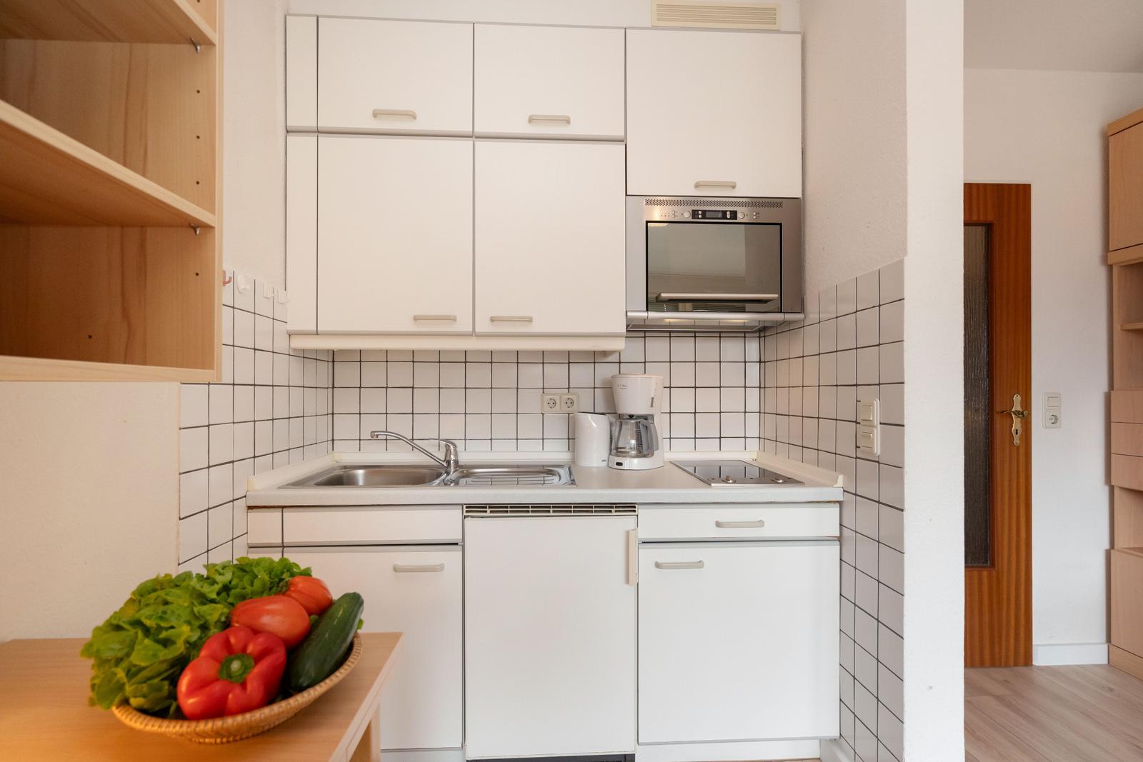 Küche mit weißen Schränken, Spüle und Kaffeemaschine. Auf dem Tisch steht eine Schale mit frischen Gemüse.