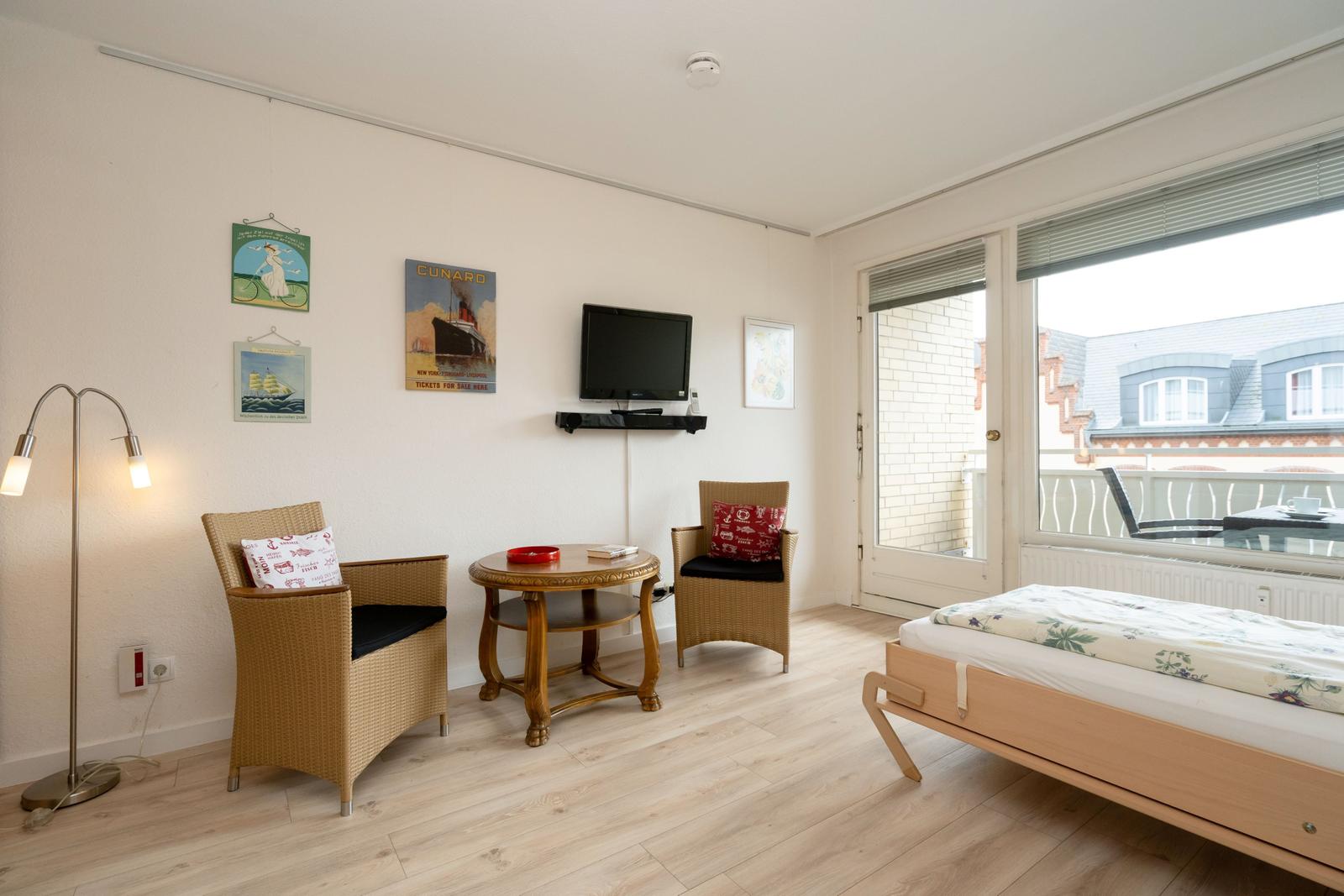 Zimmer mit Bett, zwei Stühlen, Tisch und Fernseher. Balkon mit Sicht auf Gebäude.