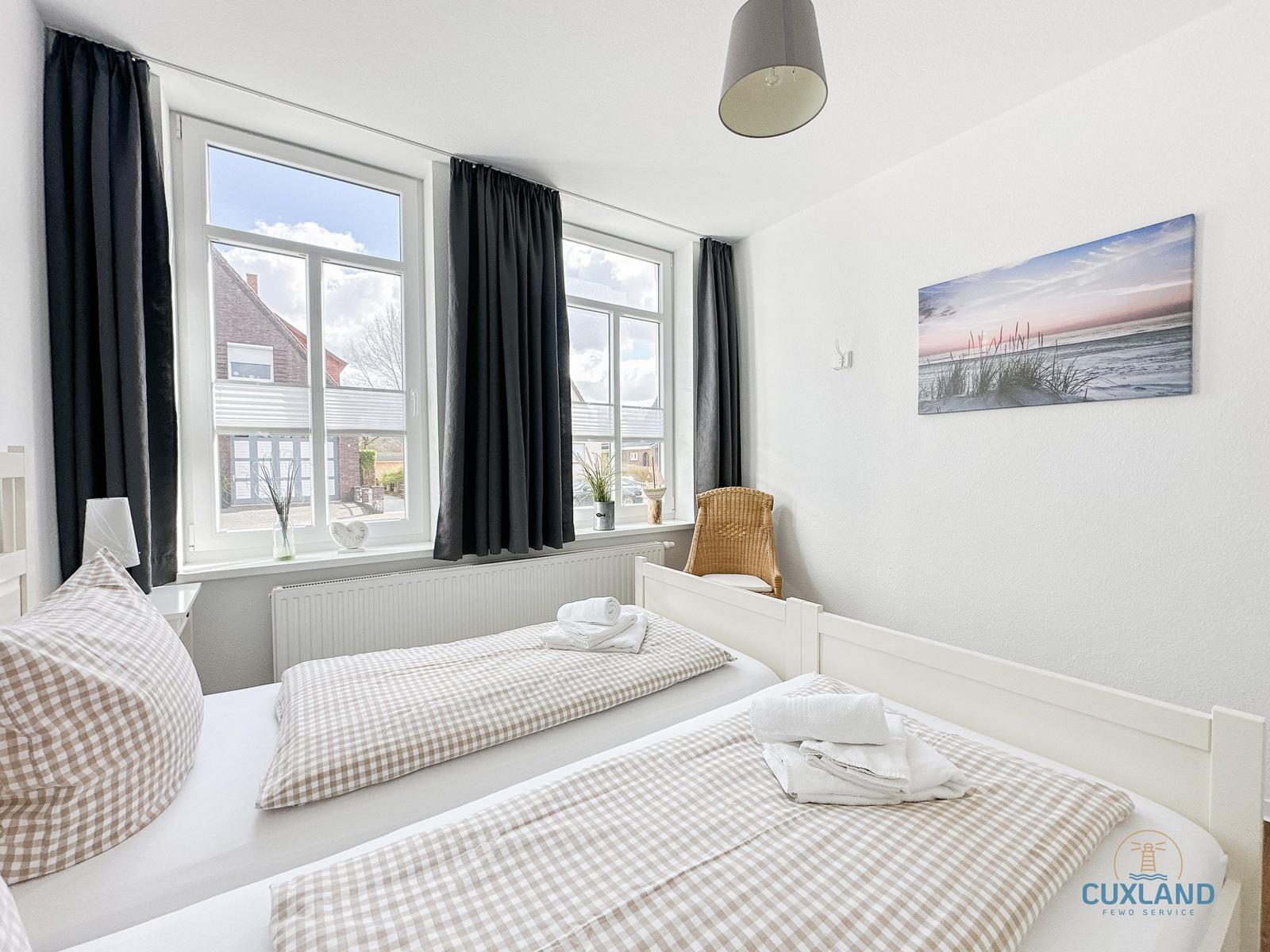 Zwei Betten mit karierten Bettwäsche, Fenster mit Blick auf Haus und Himmel, Wandbild mit Strandmotiv.