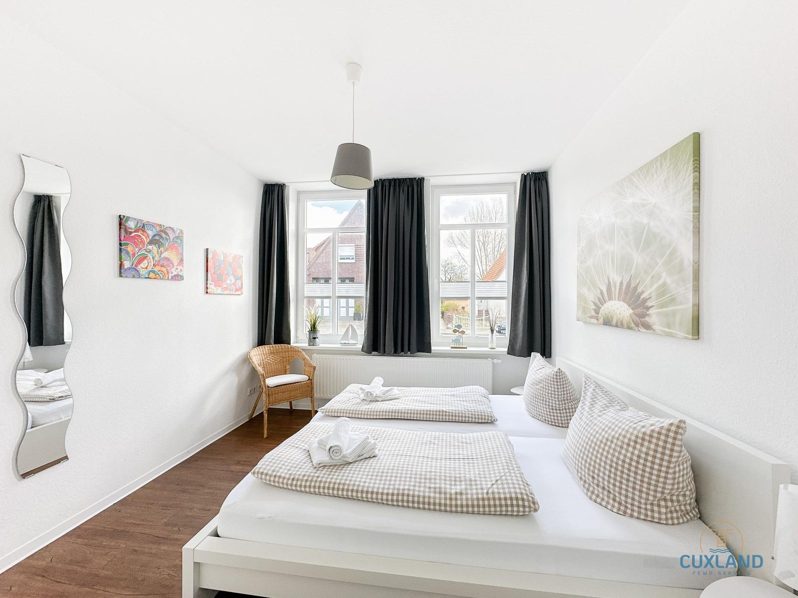 Ein Schlafzimmer mit Doppelbett, Fenster, Vorhängen und Wandbildern.