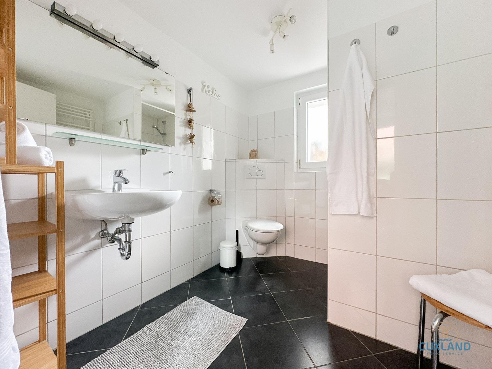 Badezimmer mit Waschbecken, Toilette und Dusche. Weiße Fliesen und schwarze Bodenfliesen.