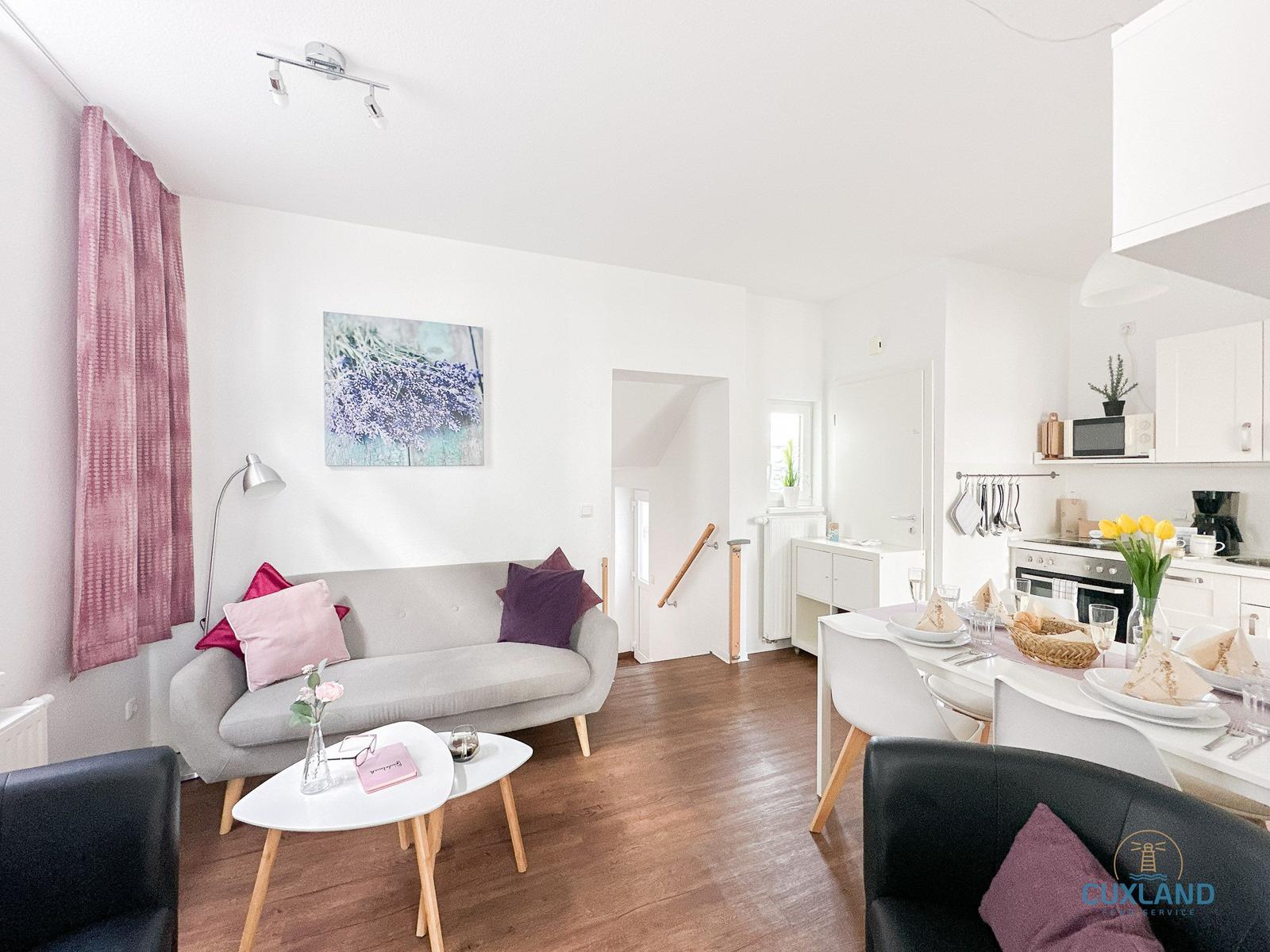 Wohn- und Essbereich mit Sofa, Tisch und Küchenzeile. Holzboden und weiße Wände.