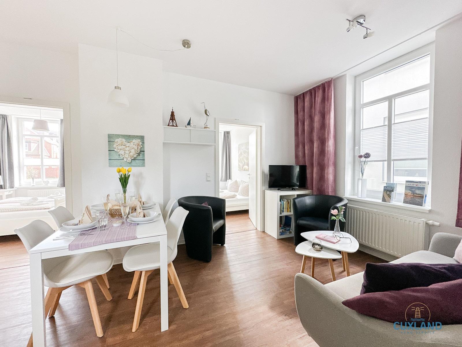 Wohn- und Essbereich mit Tisch, Stühlen, Sofa und Fernseher. Fenster mit Vorhängen und Bücherregal.
