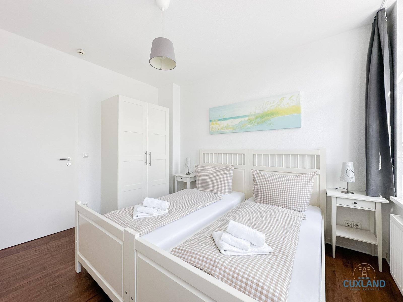 Zimmer mit Doppelbett, weißen Nachttischen und Kleiderschrank. Wandbild und graue Vorhänge.