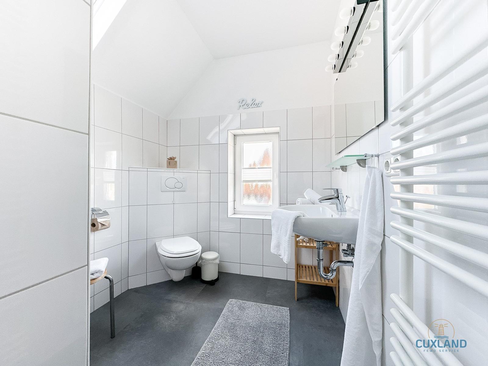Badezimmer mit Toilette, Waschbecken und Heizung. Fenster und Spiegel sichtbar.