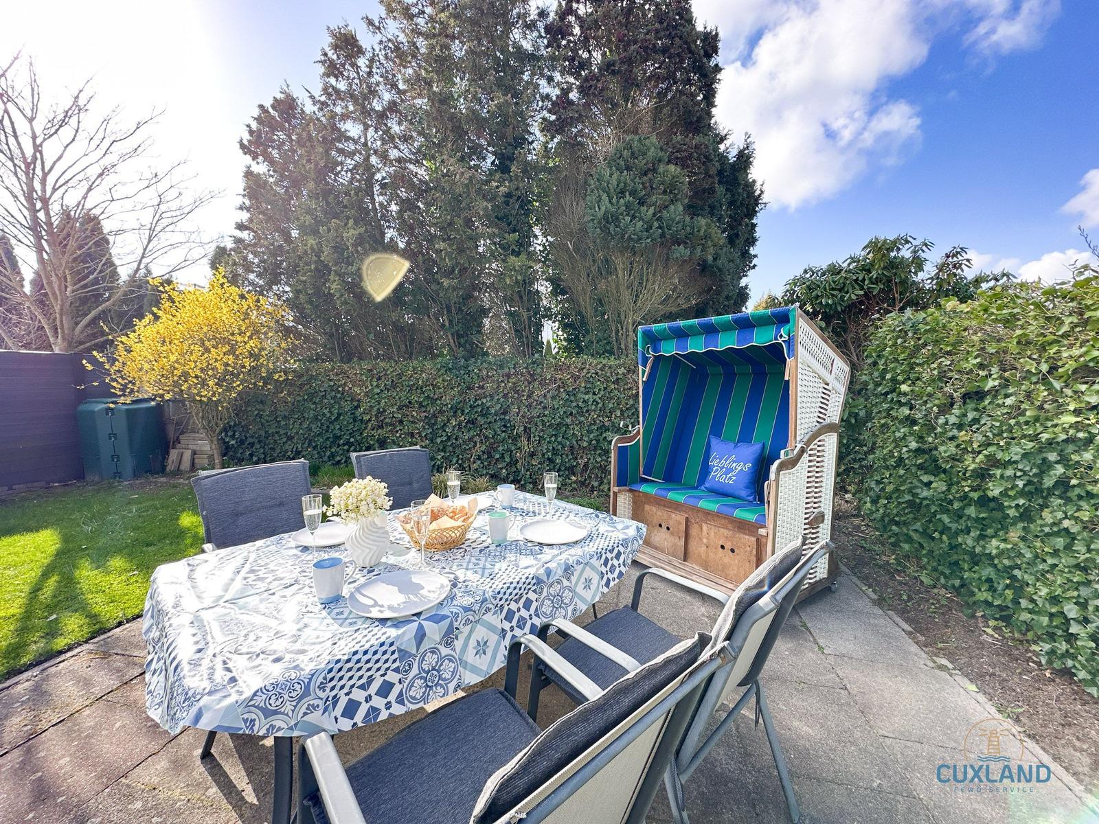 Garten mit gedecktem Tisch und Strandkorb. Hintergrund: grüne Hecke und Bäume.