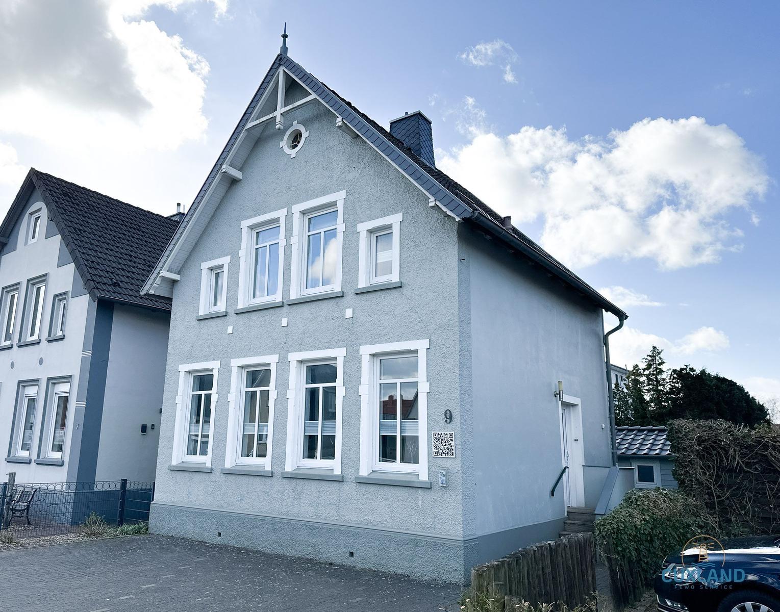 Ein zweistöckiges Haus mit grauer Fassade und weißen Fensterrahmen unter blauem Himmel.