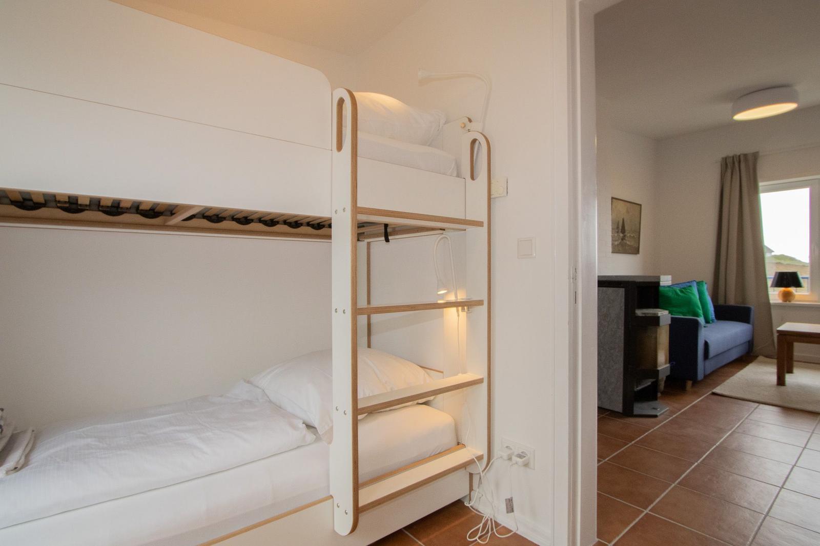 Bunk bed with white mattresses and bedside lamps. Living room with sofa in background.