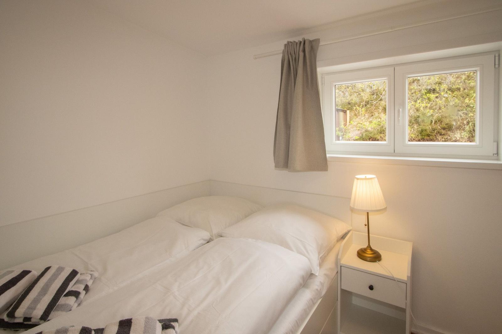 Bedroom with bed, nightstand, and lamp. Window with view of greenery.