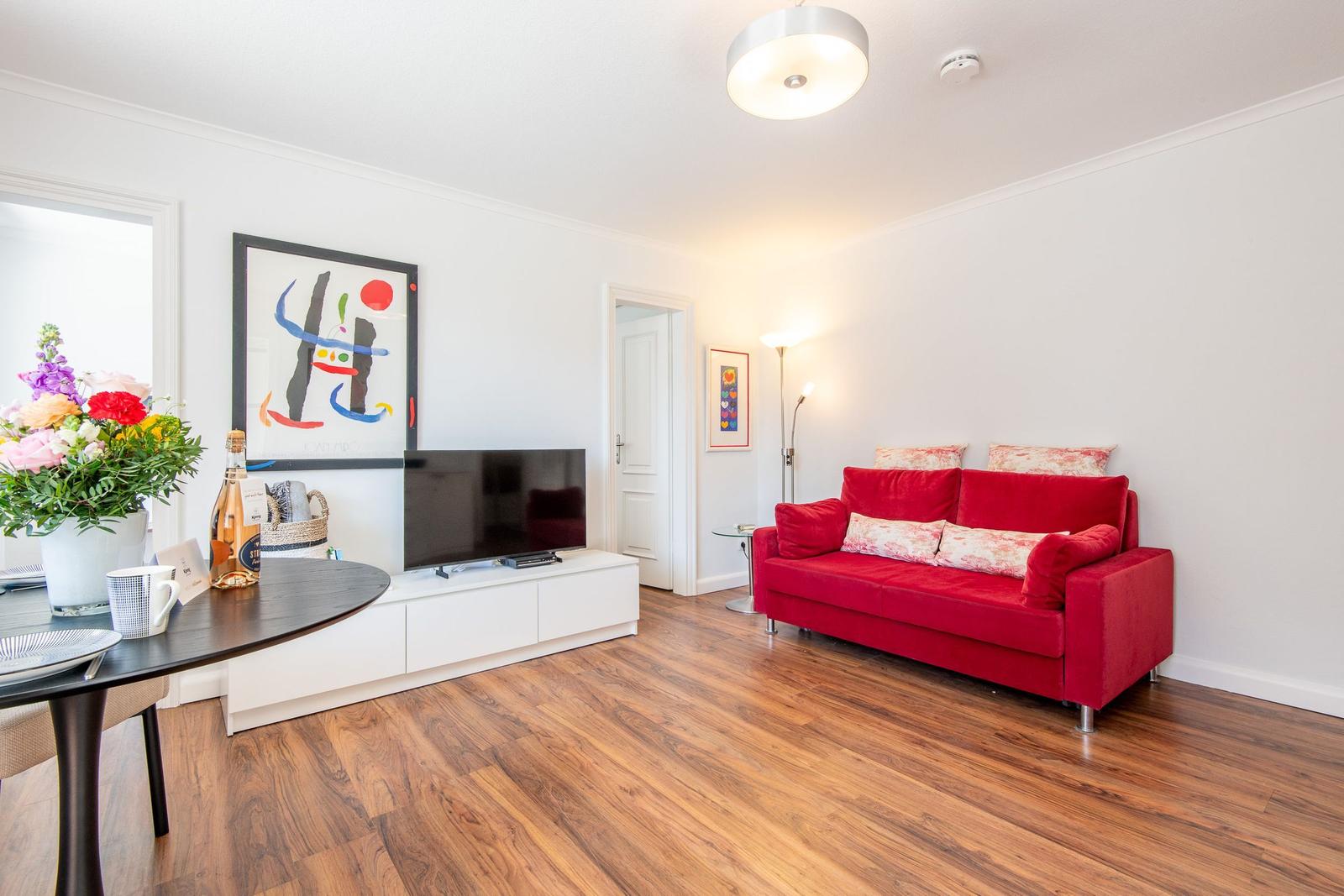 A living room with red sofa, wooden floor, and TV on white cabinet.