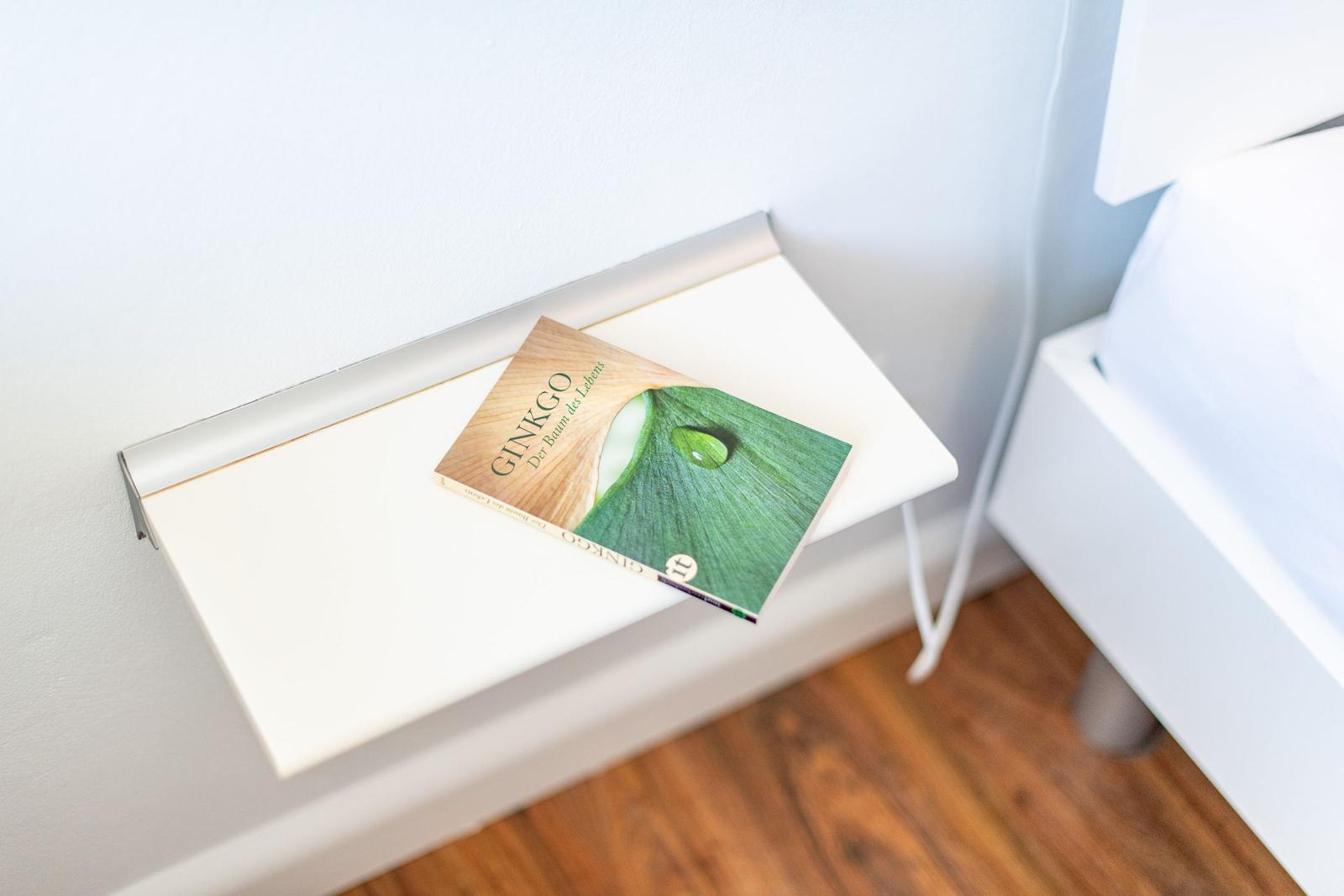A book rests on a white bedside table next to a bed.