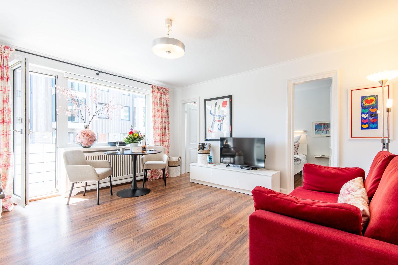 Living room with red sofa, TV and table with chairs. Passage to bedroom.