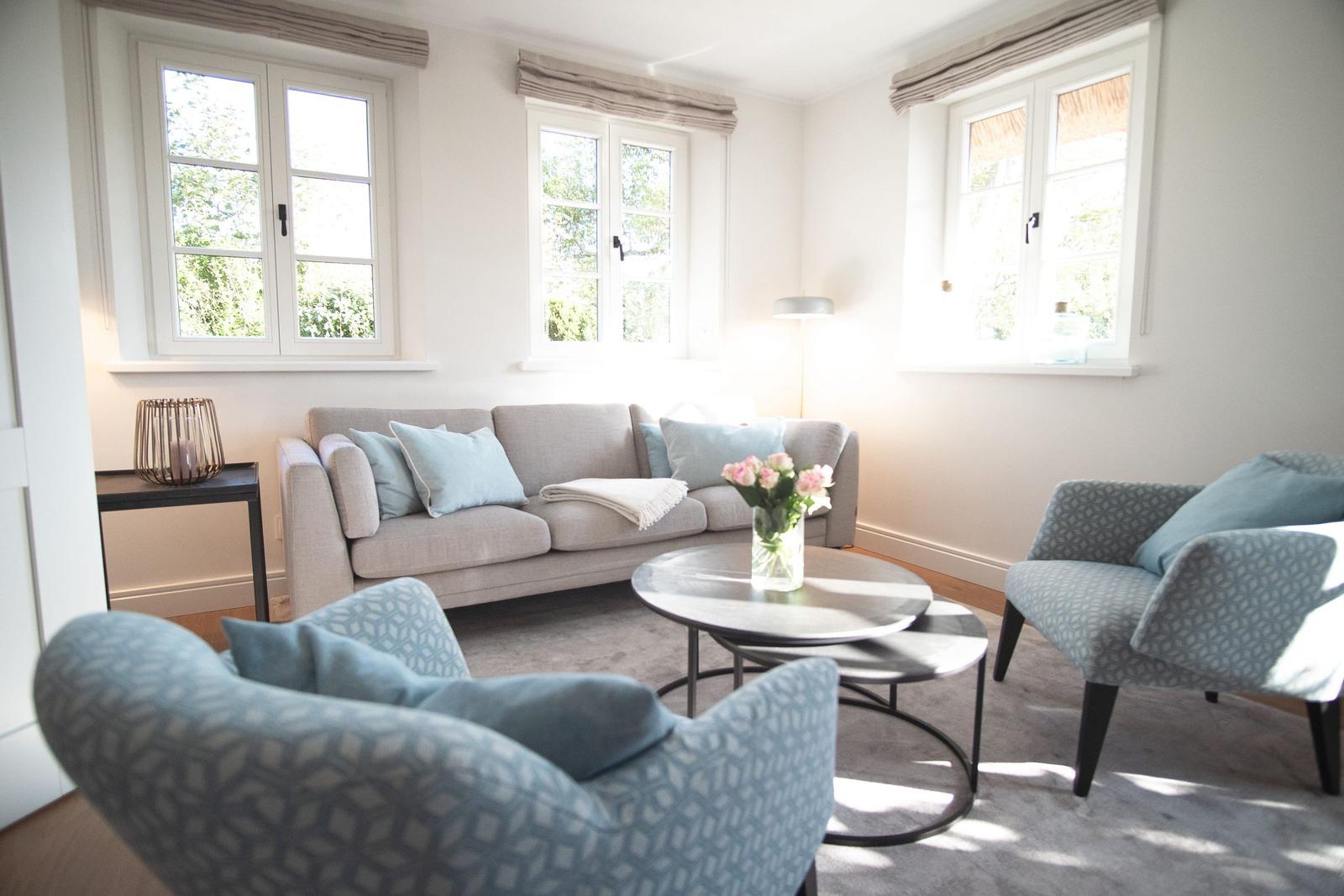 Wohnzimmer mit Sofa, zwei Sesseln und runden Tischen. Drei Fenster mit Blick auf Grün.
