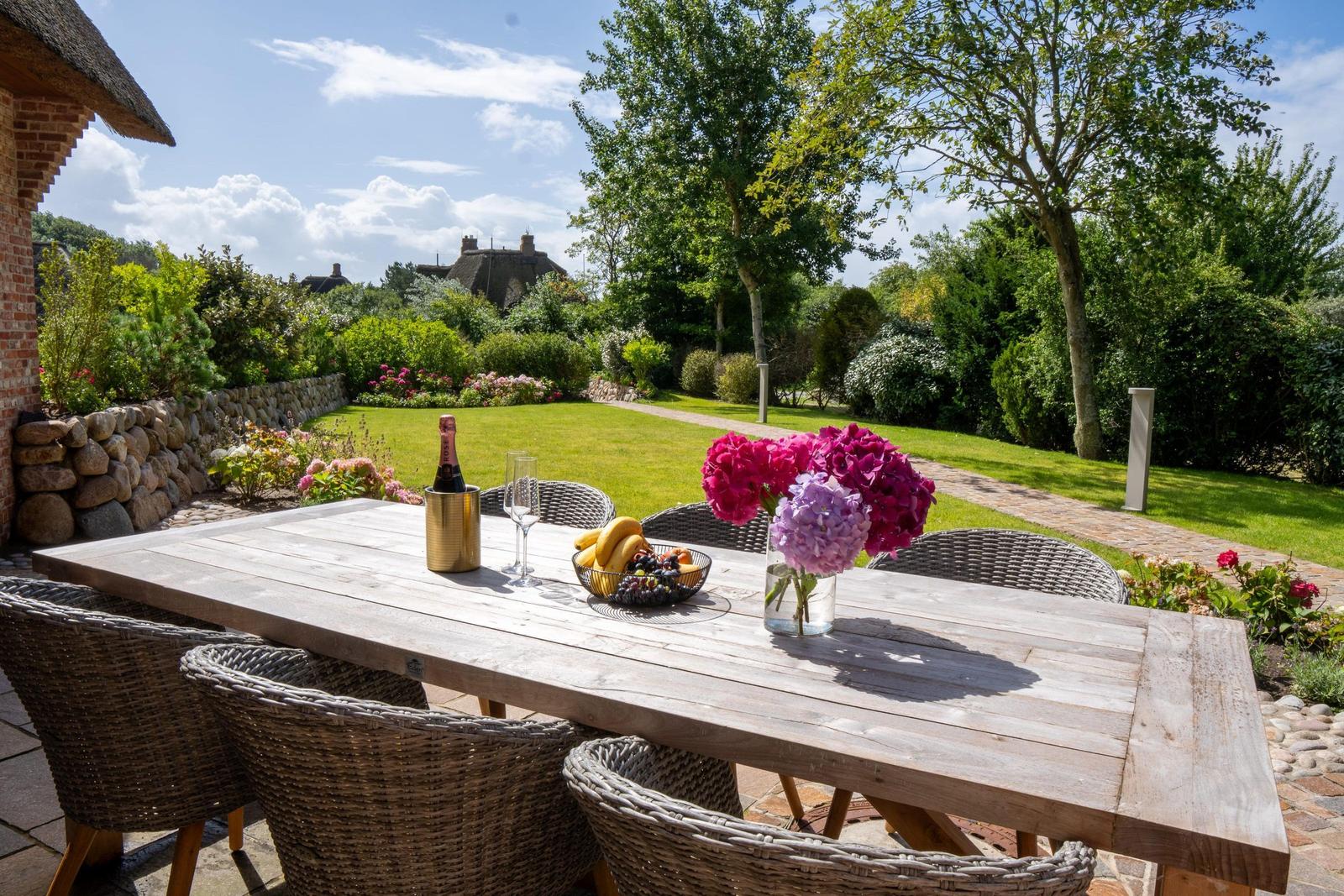 Holztisch mit Stühlen, Blumen und Obst auf Terrasse mit Gartenblick.