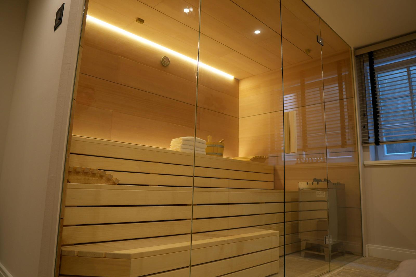 Sauna mit Holzbank und Glaswand. Dampfheizung und Fenster mit Jalousie.