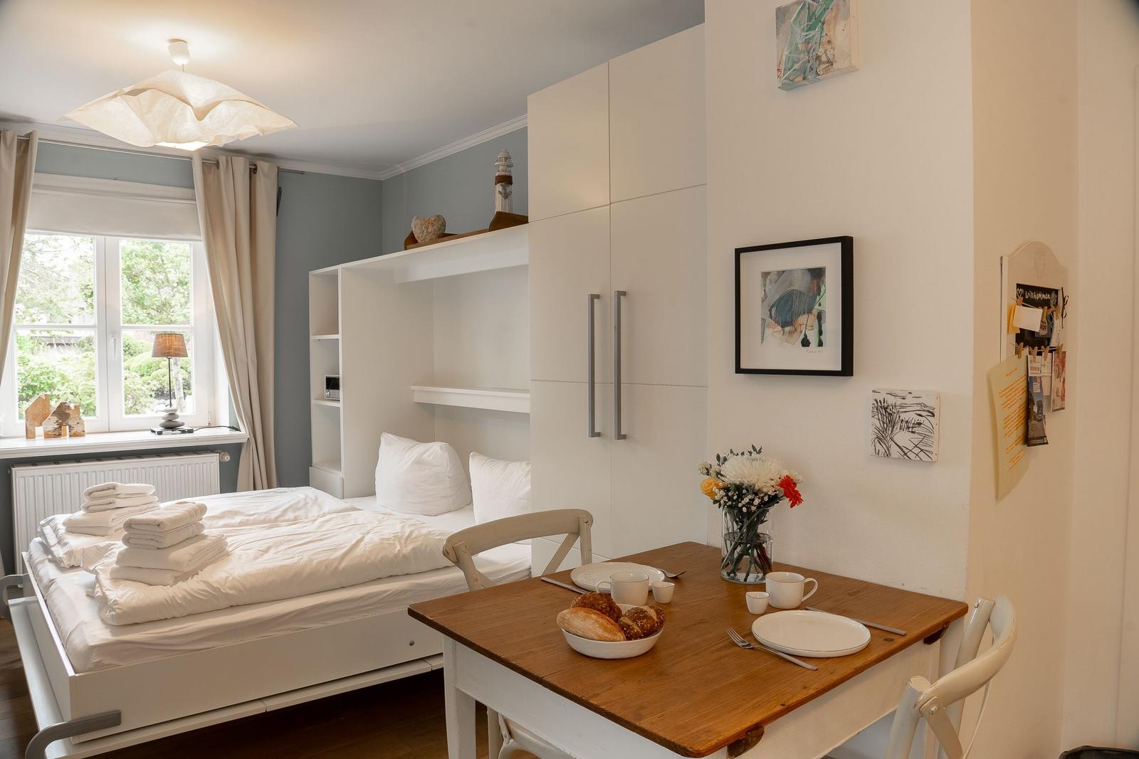 Schlafzimmer mit Bett, Tisch und Fenster. Auf dem Tisch stehen Tassen und Brot.