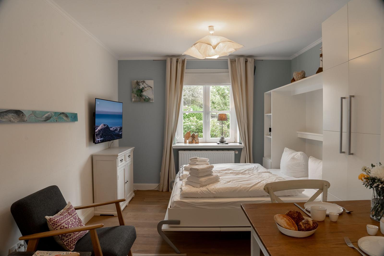 Schlafzimmer mit Bett, TV und Essplatz. Fenster mit Blick auf Grün.
