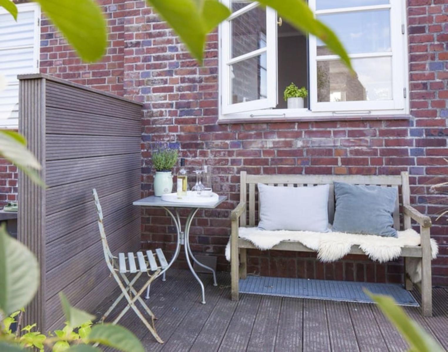 Terrasse mit Holzbank, Kissen und Tisch vor Backsteinmauer.