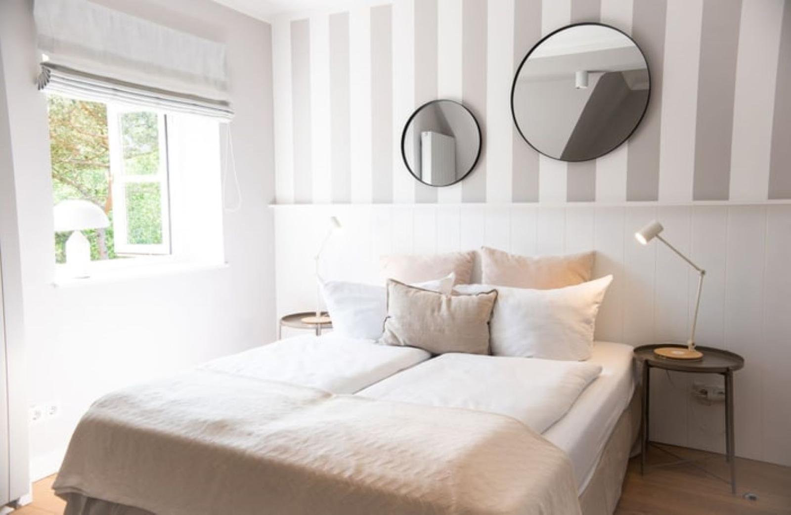 Schlafzimmer mit Bett, zwei Nachttischen und runden Spiegeln an gestreifter Wand.