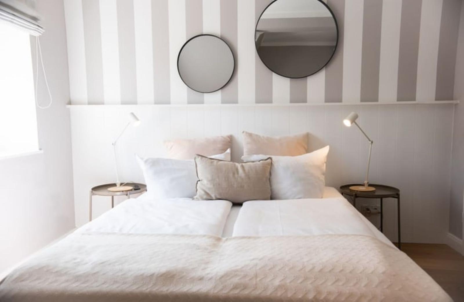 Schlafzimmer mit gestreifter Wand, zwei runden Spiegeln und einem Bett mit Kissen.