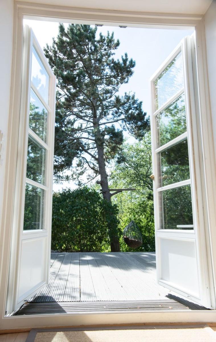 Durch offene Fenster blickt man auf eine Terrasse mit Hängesessel und einen hohen Baum.