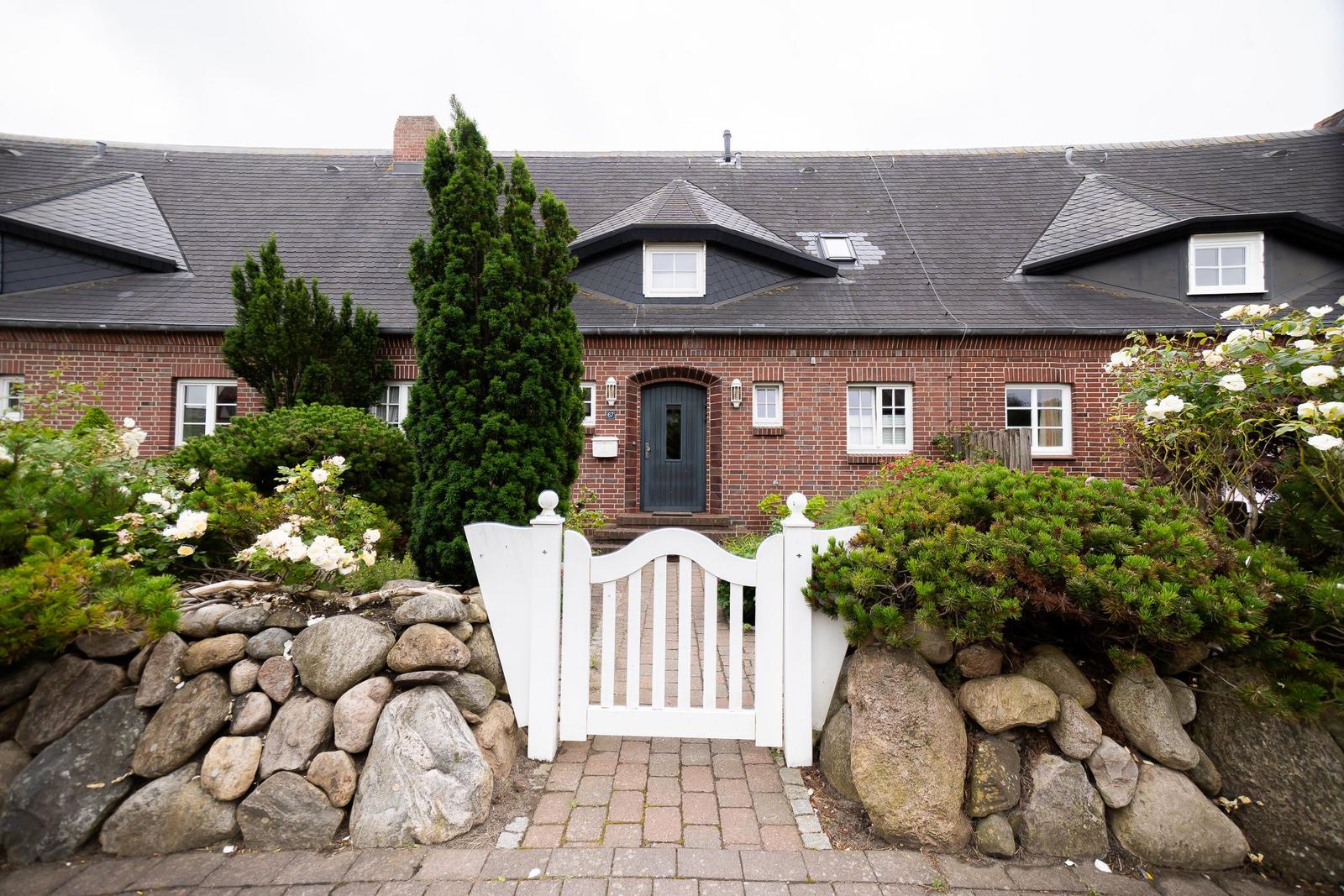Red brick building with white door and garden gate. Surrounded by shrubs and stones.