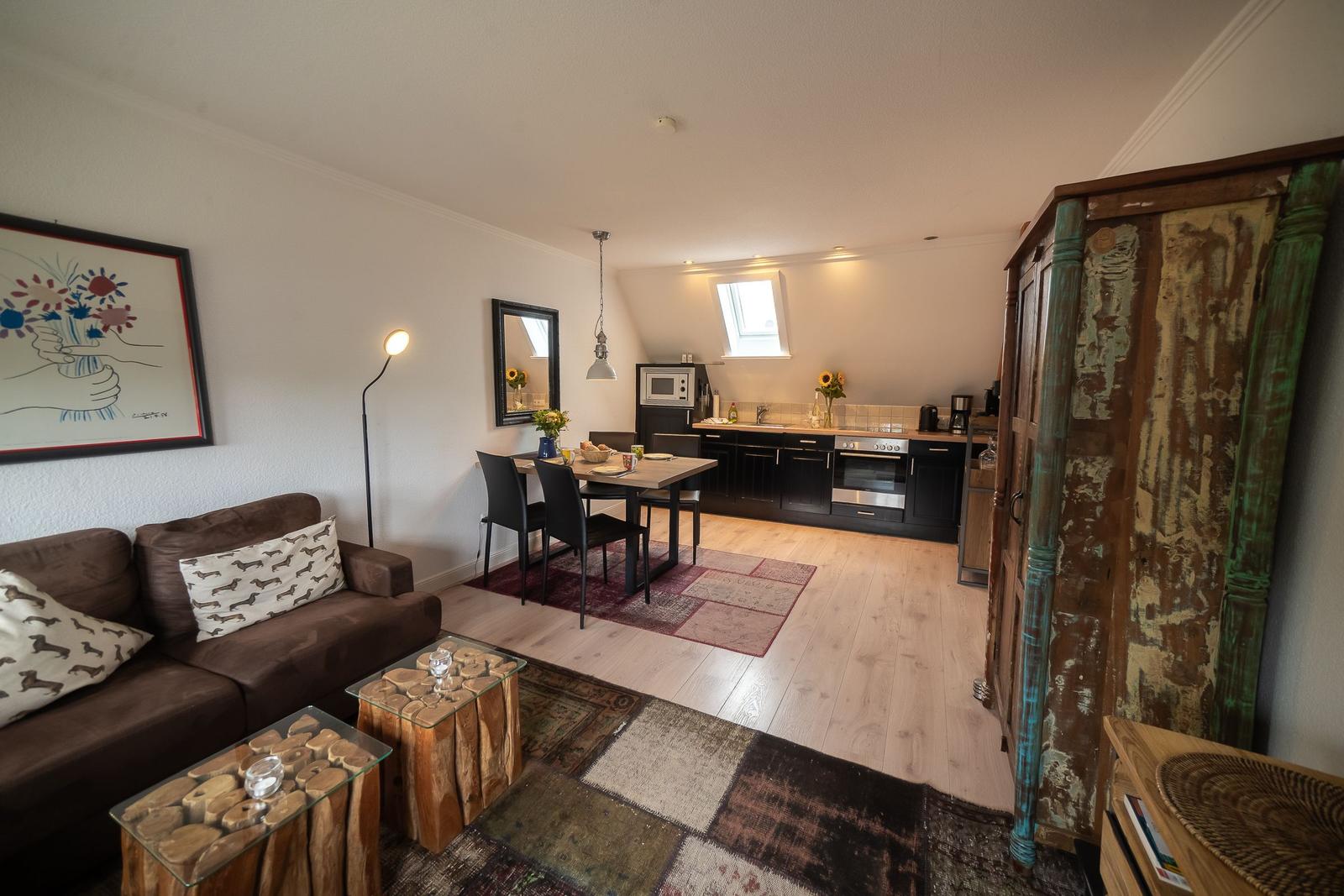 Living and dining area with kitchen under sloped ceiling. Wooden floor, sofa, table, and cabinet visible.