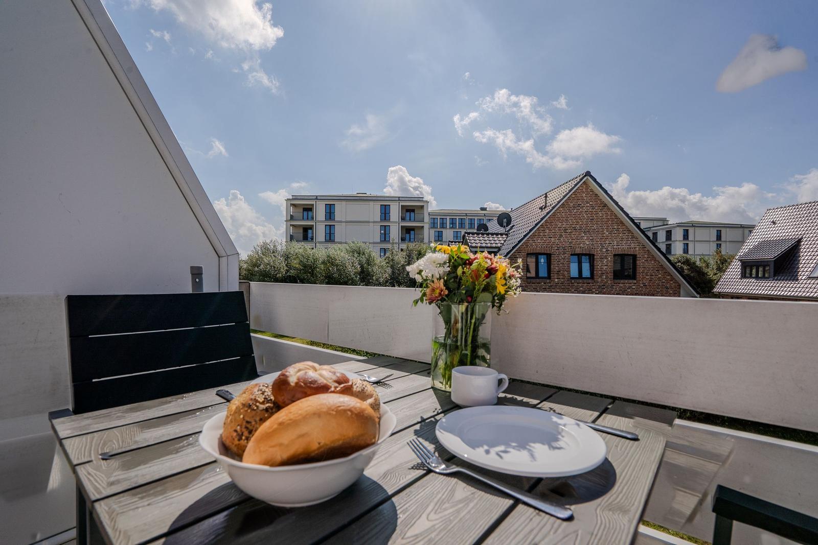 Terrasse mit Tisch, Stühlen, Brot und Blumen. Blick auf Wohnhäuser und Himmel.