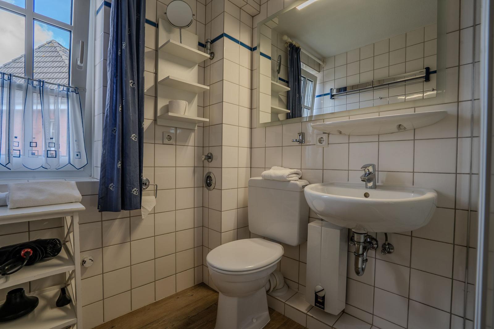 Badezimmer mit Toilette, Waschbecken und Dusche. Fenster mit Vorhängen.
