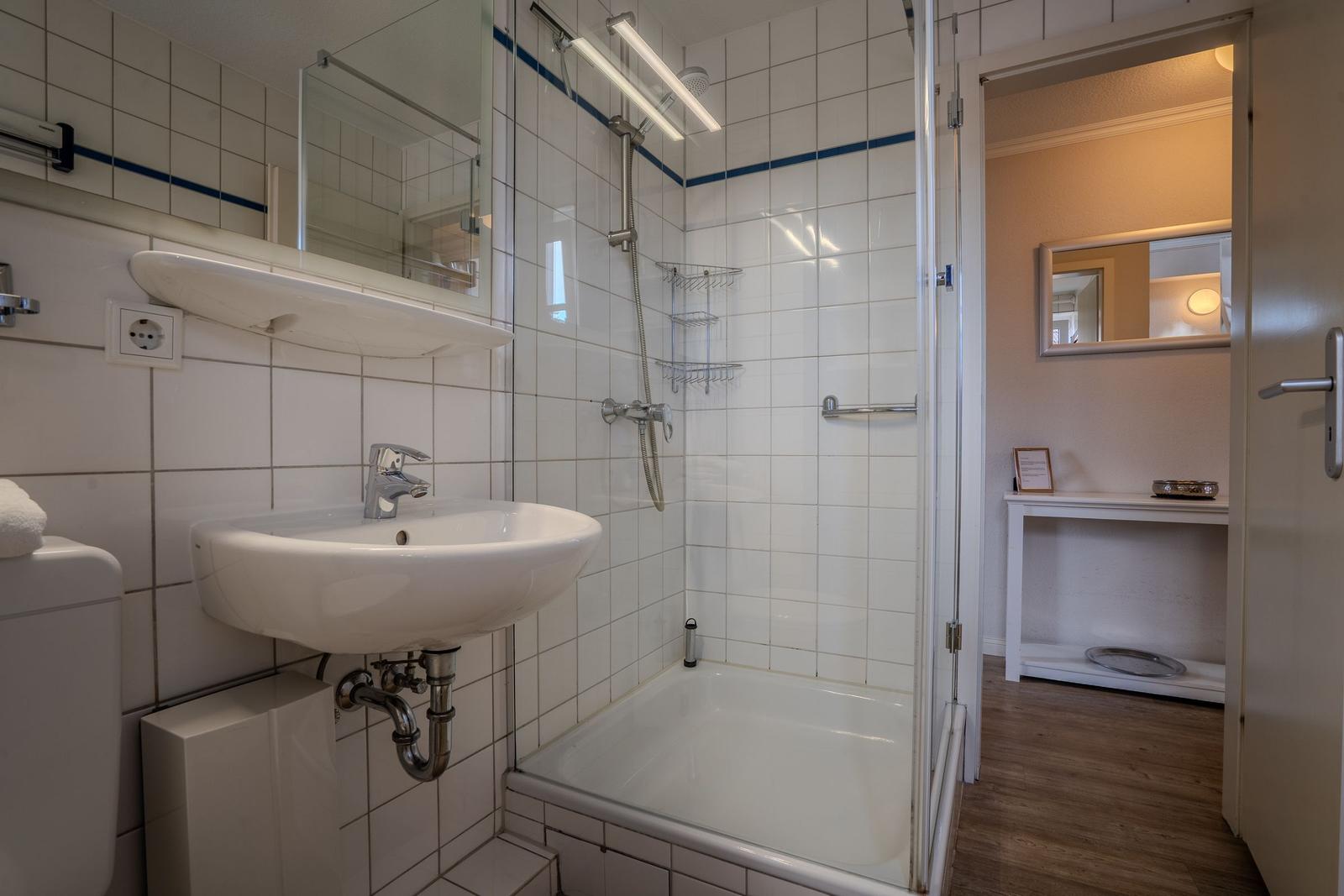 Bad mit Dusche, Waschbecken und Spiegel. Tür führt in einen Raum mit Schreibtisch.