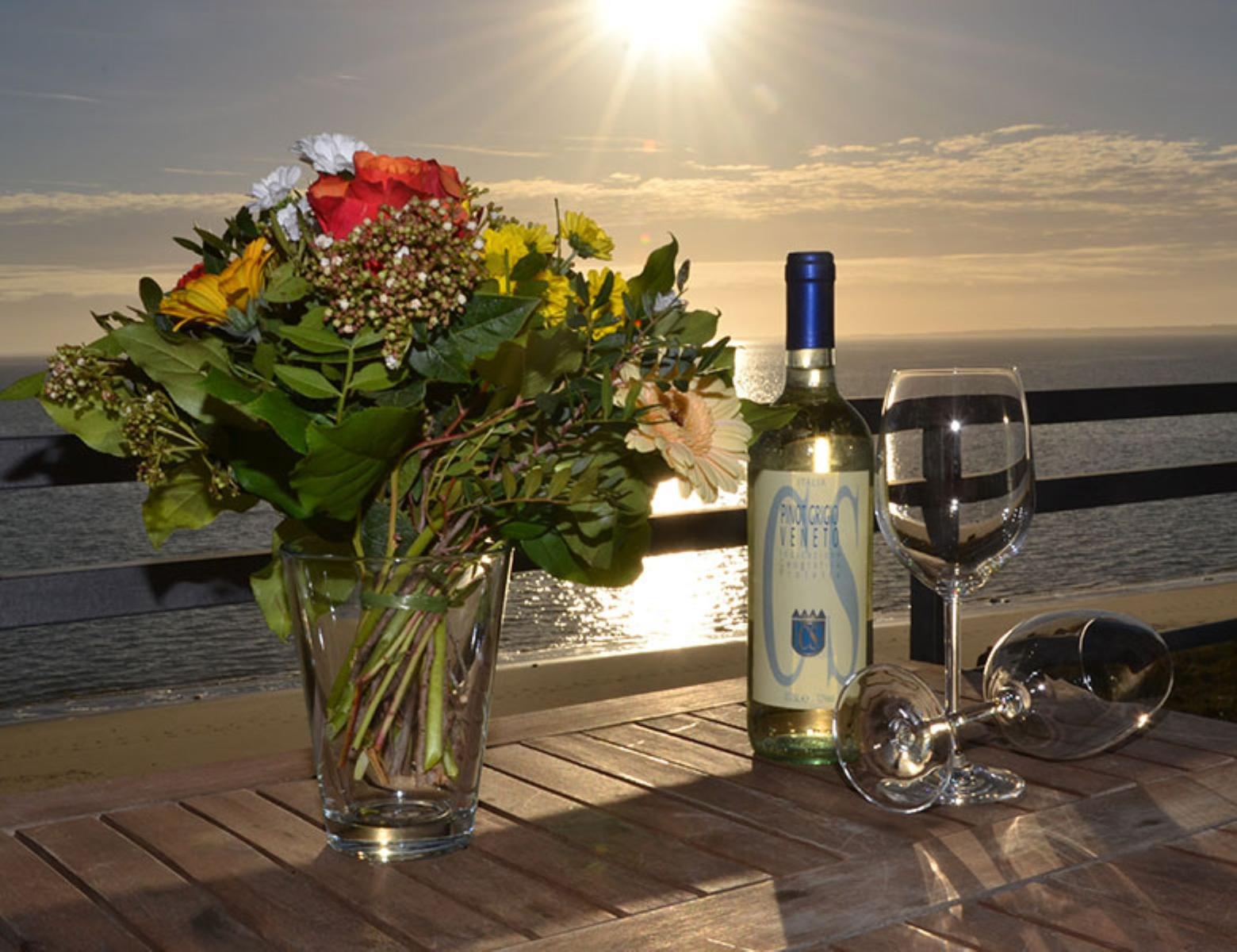 Auf der Terrasse steht ein Tisch mit Blumen, Wein und Gläsern vor Sonnenuntergang.