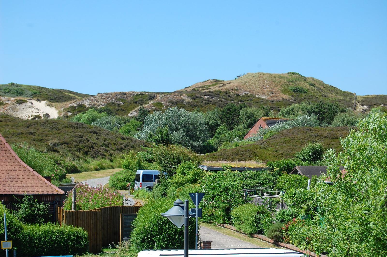 Häuser und Grünflächen unter blauem Himmel mit Hügeln im Hintergrund.