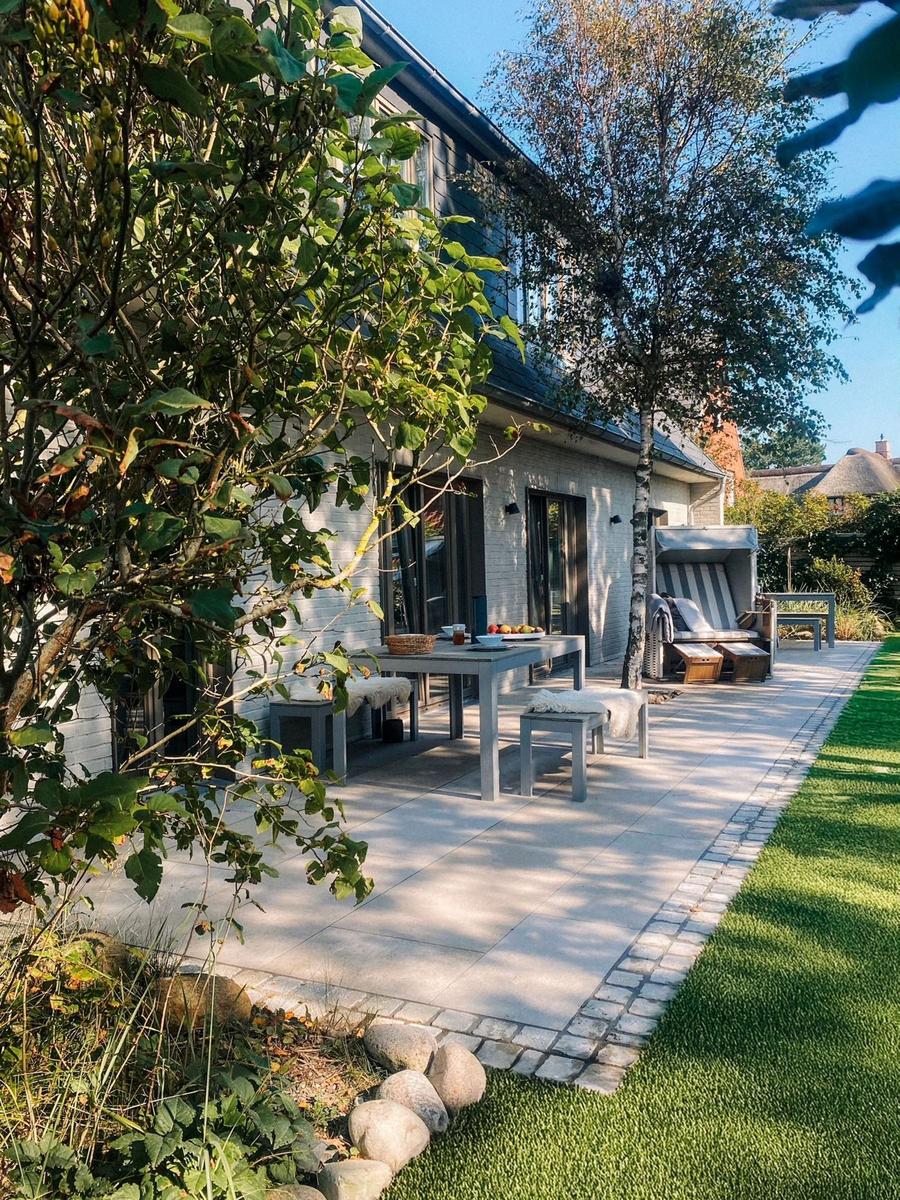 Terrasse mit Tisch, Stühlen und Liege unter Bäumen. Grünfläche und Steinweg.