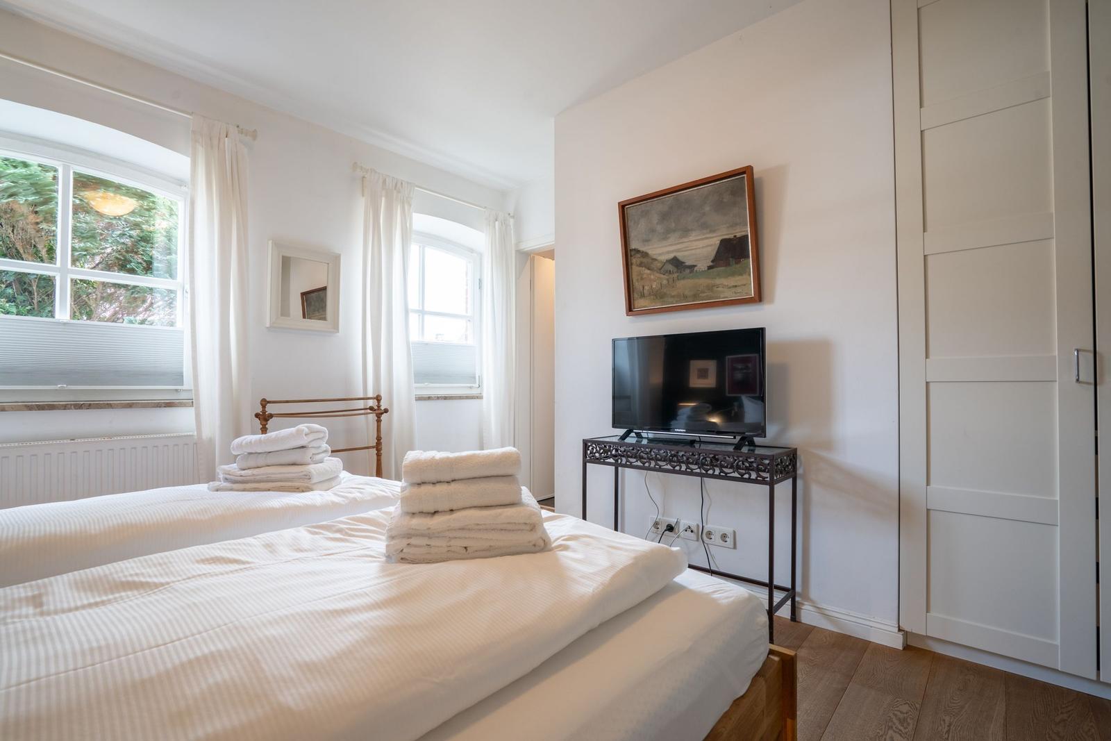 Two beds with white sheets, windows with white curtains, and TV on table.