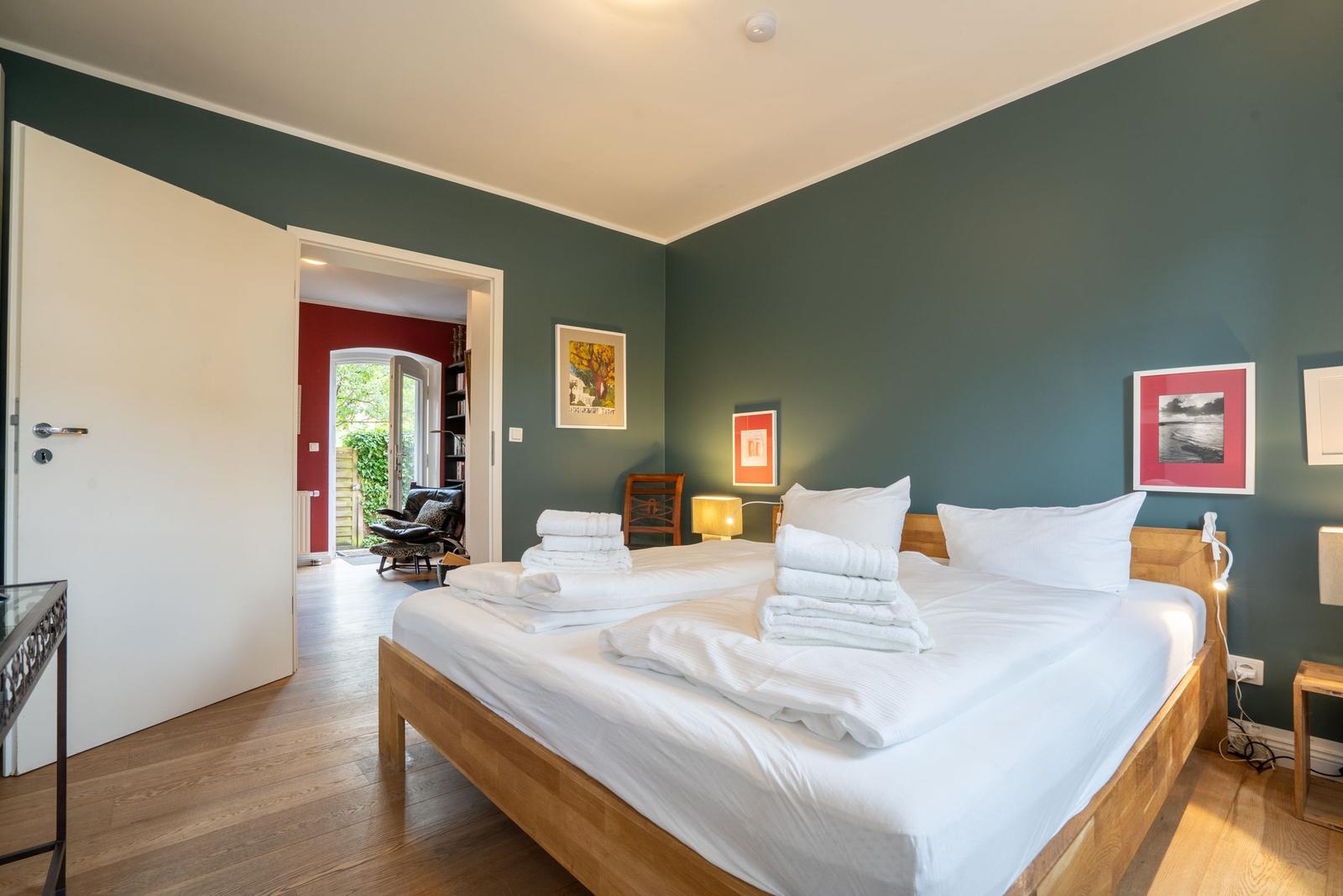 Bedroom with wooden bed, white linens, and green walls. Door leads to living area with red wall.