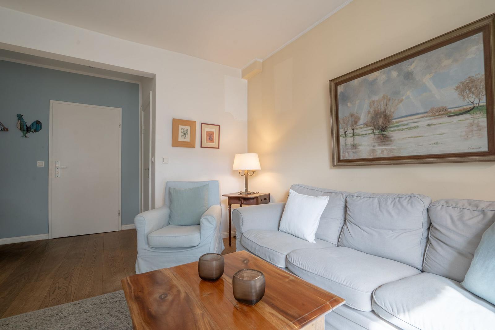 Living room with sofa, armchair, and wooden table. Wall painting and lamp visible.
