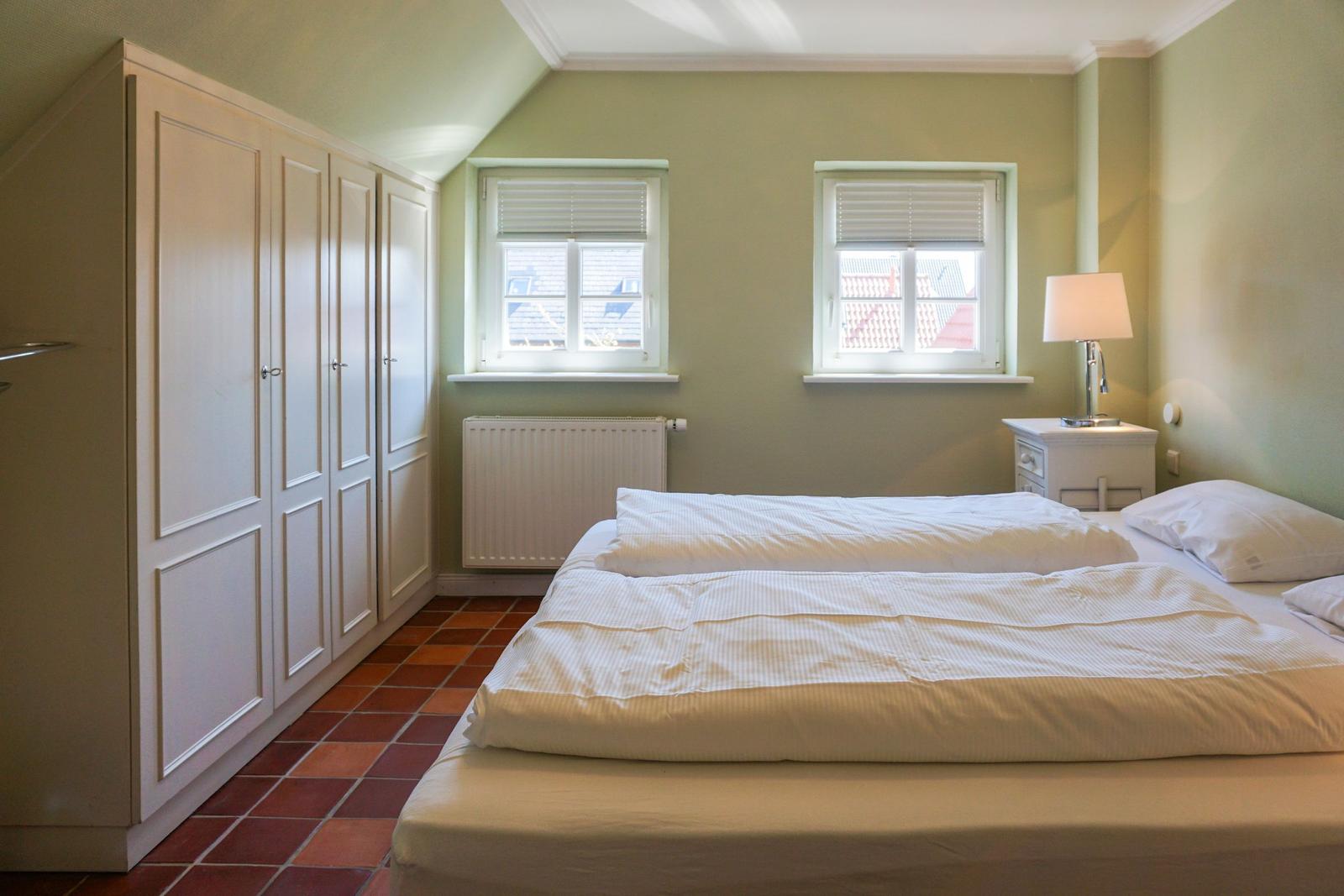 Schlafzimmer mit Bett, Kleiderschrank und zwei Fenstern.
