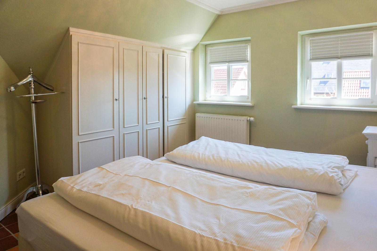 Schlafzimmer mit Doppelbett, Kleiderschrank und zwei Fenstern.