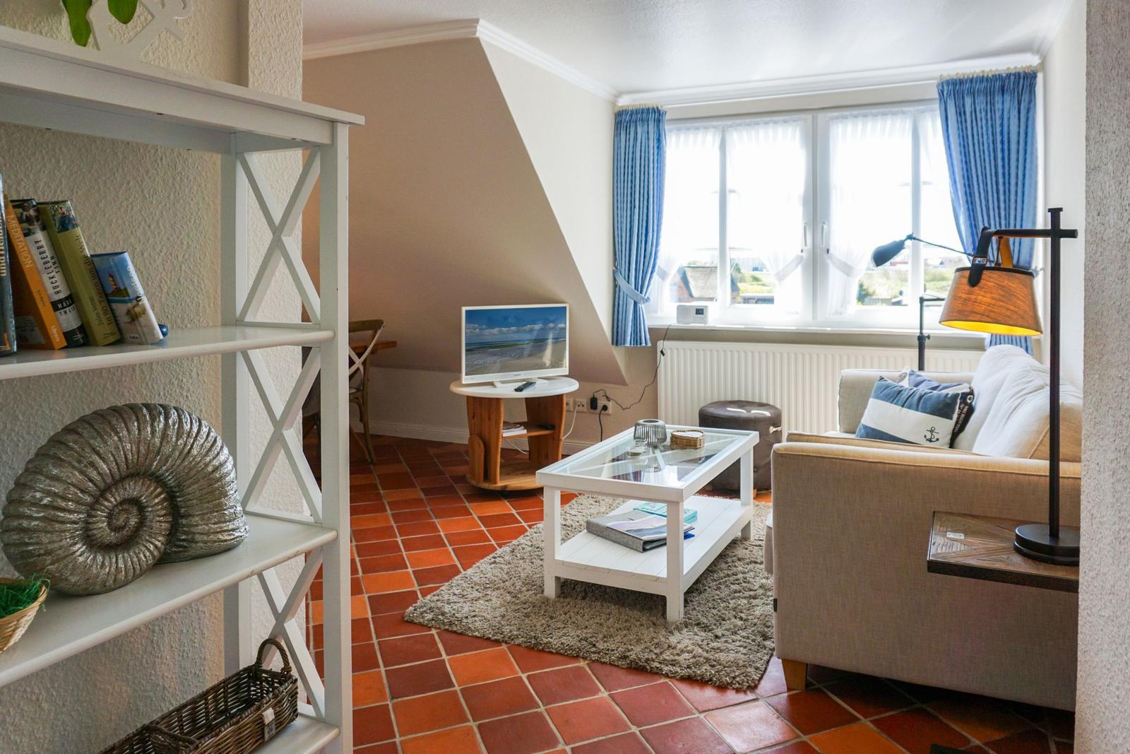 Wohnzimmer mit Sofa, Glas-Tisch und TV unter Dachschräge. Terrakottaboden und Bücherregal.
