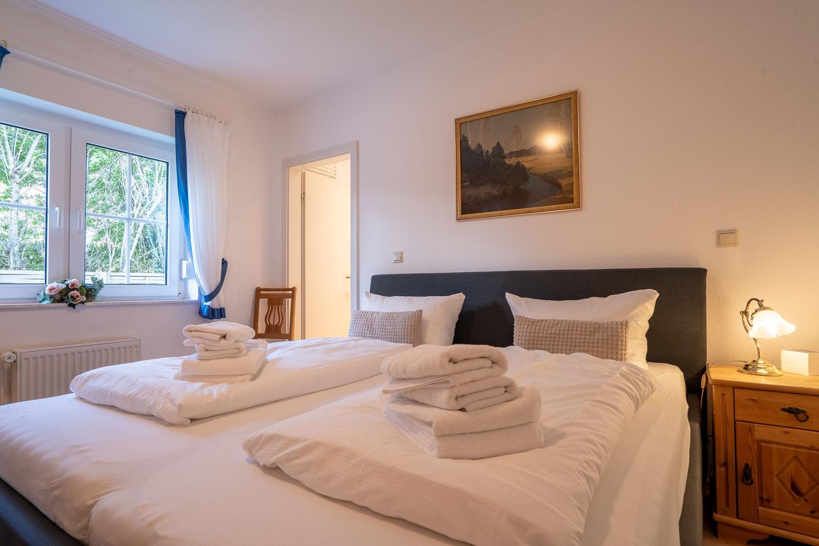 Double bed with white linens and folded towels. Window with blue curtains and nightstand with lamp.