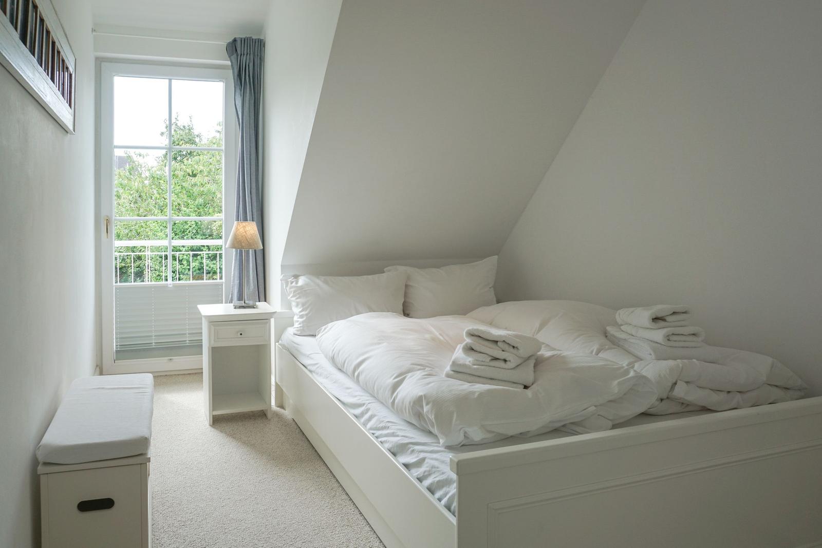 Bedroom with sloped ceiling, white bed, and window to balcony.