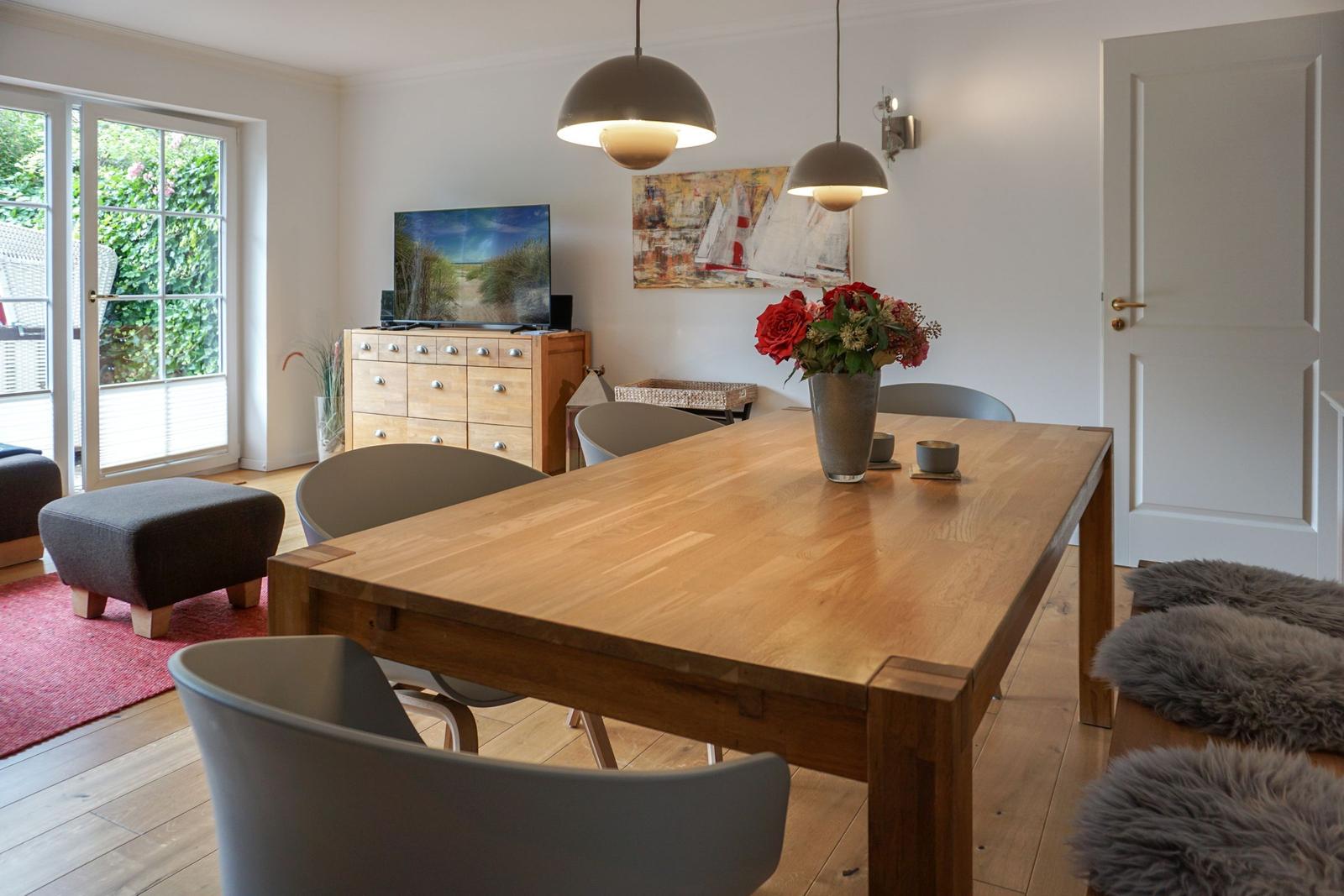 Dining room with wooden table, chairs, and flower arrangement. Windows and door lead outside.