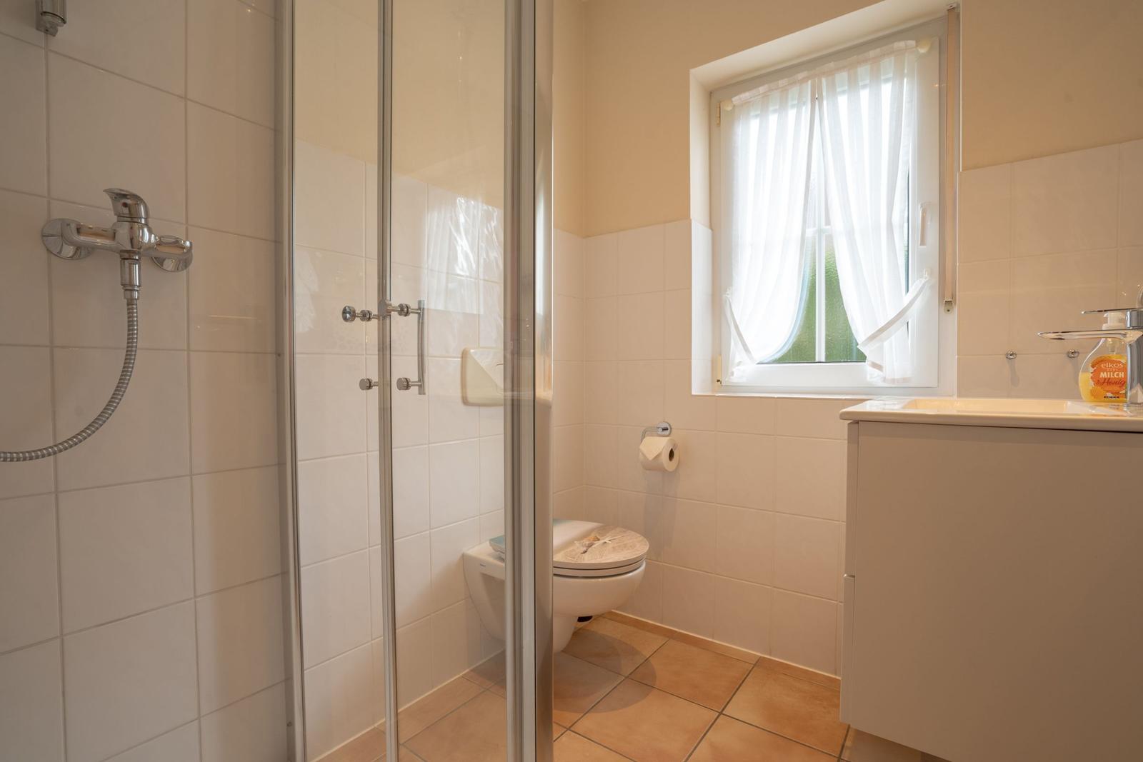 Bathroom with shower, toilet, and sink. Window with white curtains.