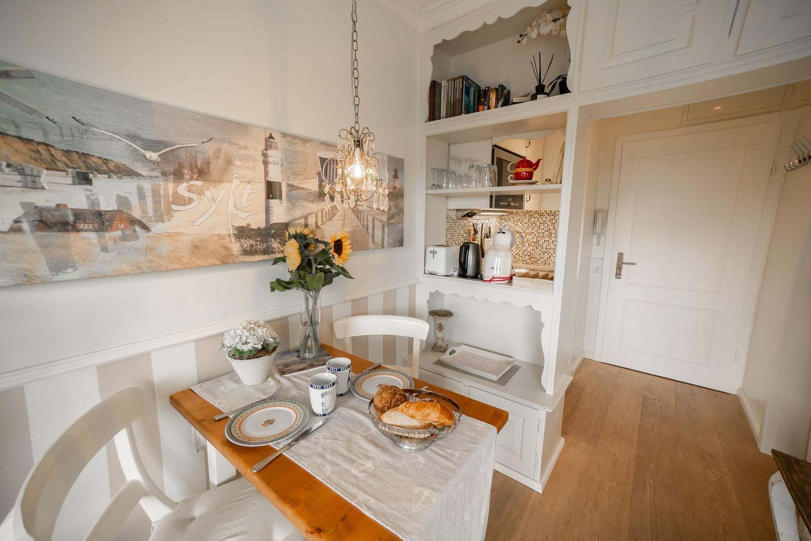 Dining table with tablecloth, mugs, and bread. Kitchenette and wall art with pendant light.