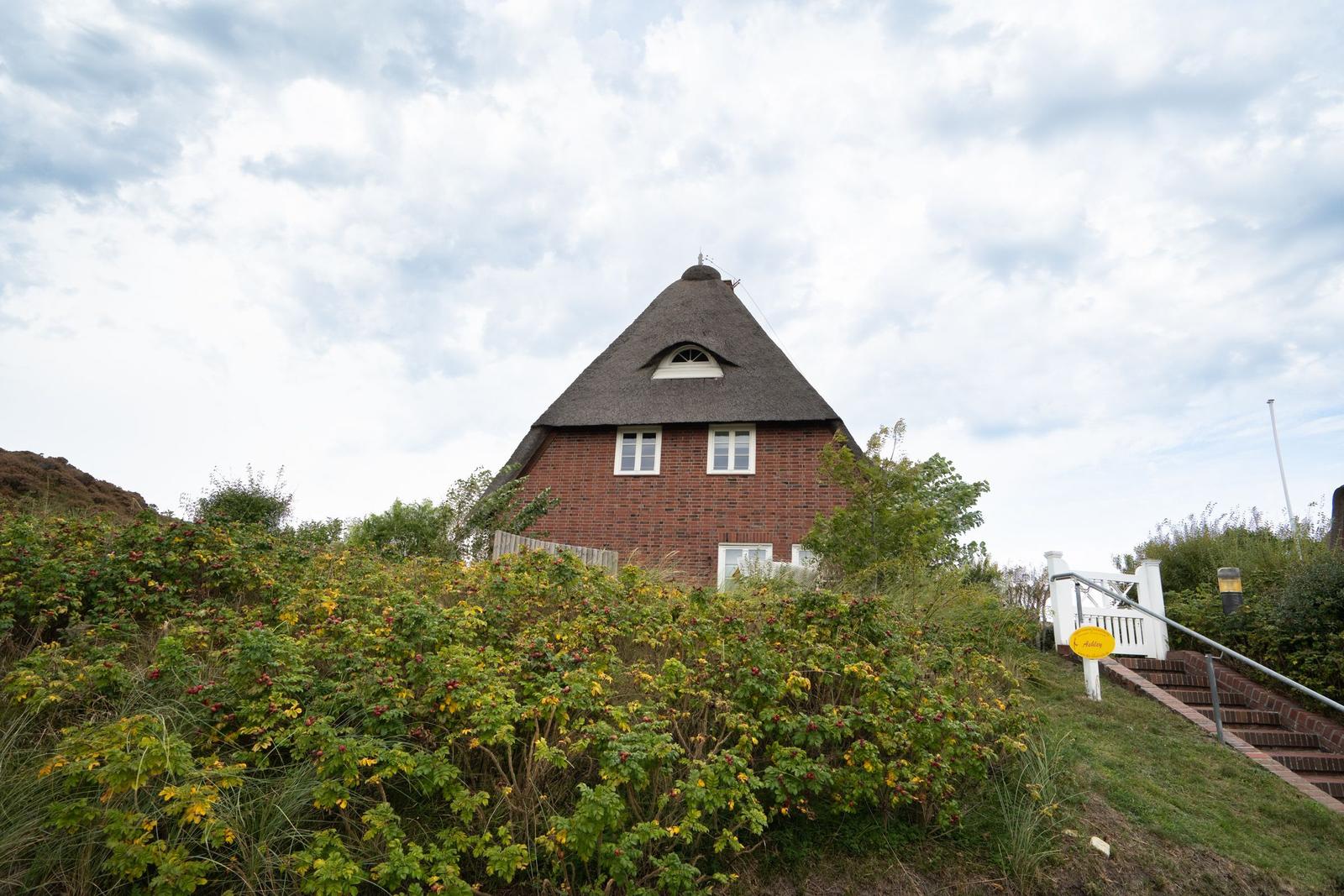 Rotes Backsteingebäude mit Strohdach und weißen Fenstern. Umgeben von grüner Vegetation und einer Treppe.