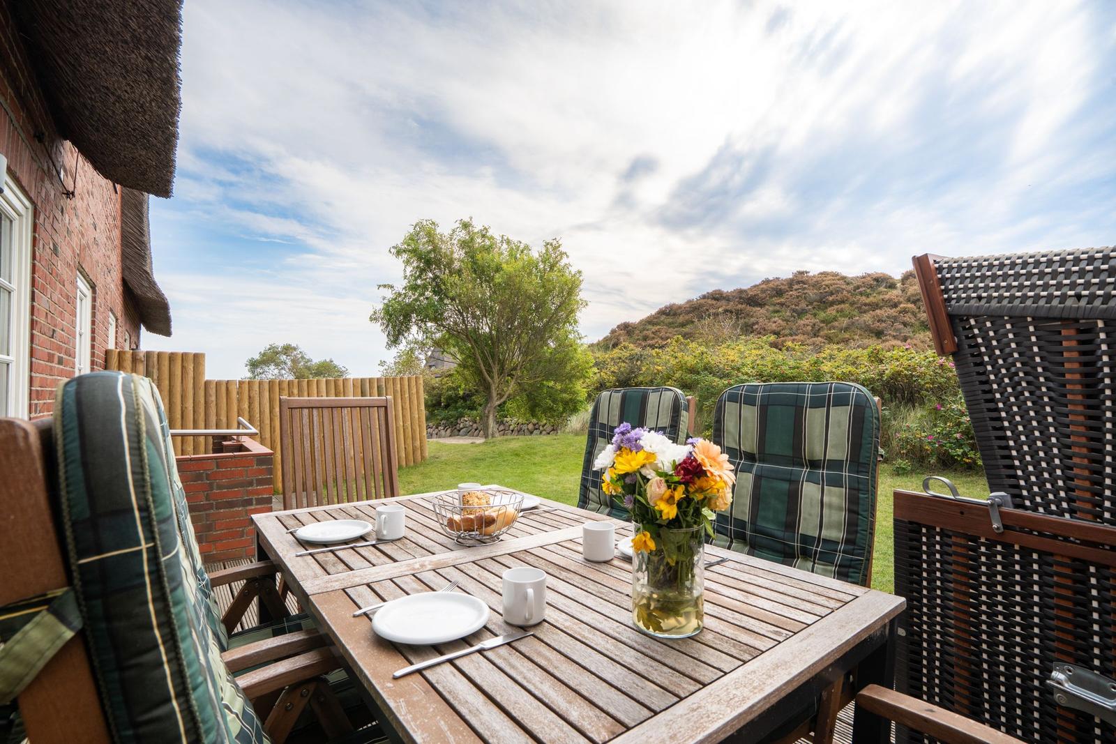 Terrasse mit Holztisch, Stühlen und Blumen. Blick auf grünen Garten und Hügel.