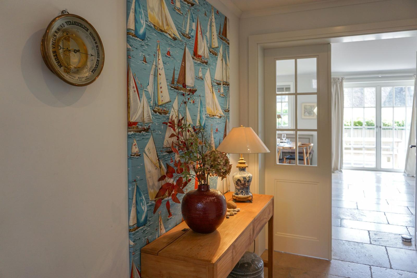Entryway with sailboat wallpaper, wooden console, vase, and barometer.