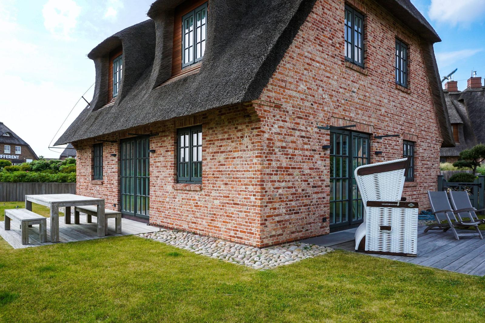 Red brick house with thatched roof, garden with table and lounge chairs.