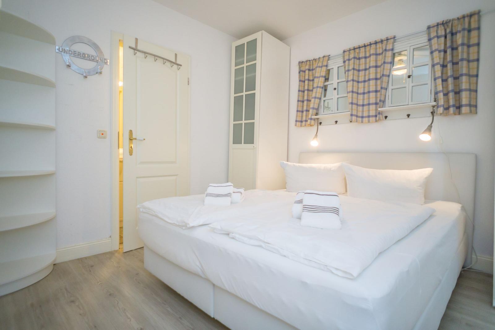 Schlafzimmer mit weißem Bett, Gardinen und Schrank. Tür führt in Bad.