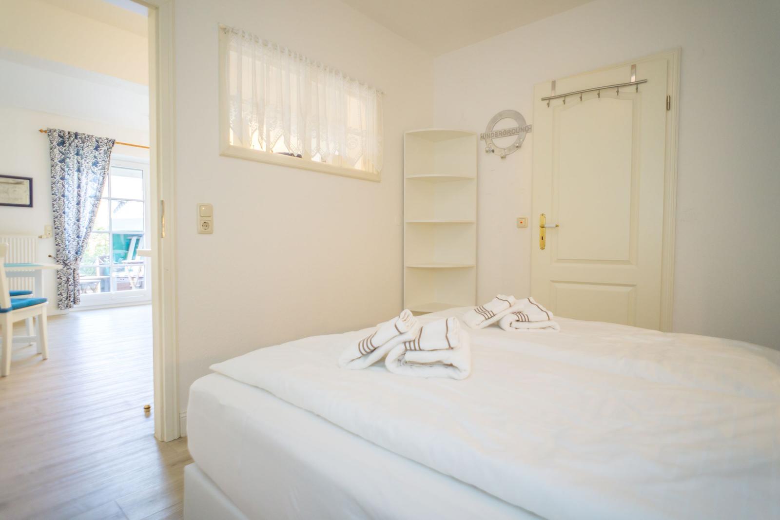 Schlafzimmer mit Bett, Handtuch und Tür. Durchgang zu Wohnbereich mit Tisch und Stuhl.